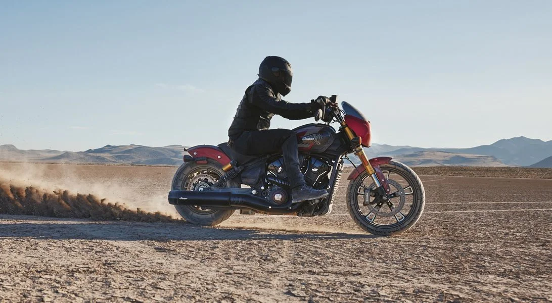 A rider in full gear leans a red Indian Scout through a hard turn on a dry desert lakebed, kicking up a wall of dust — hands on the throttle, riding forward.